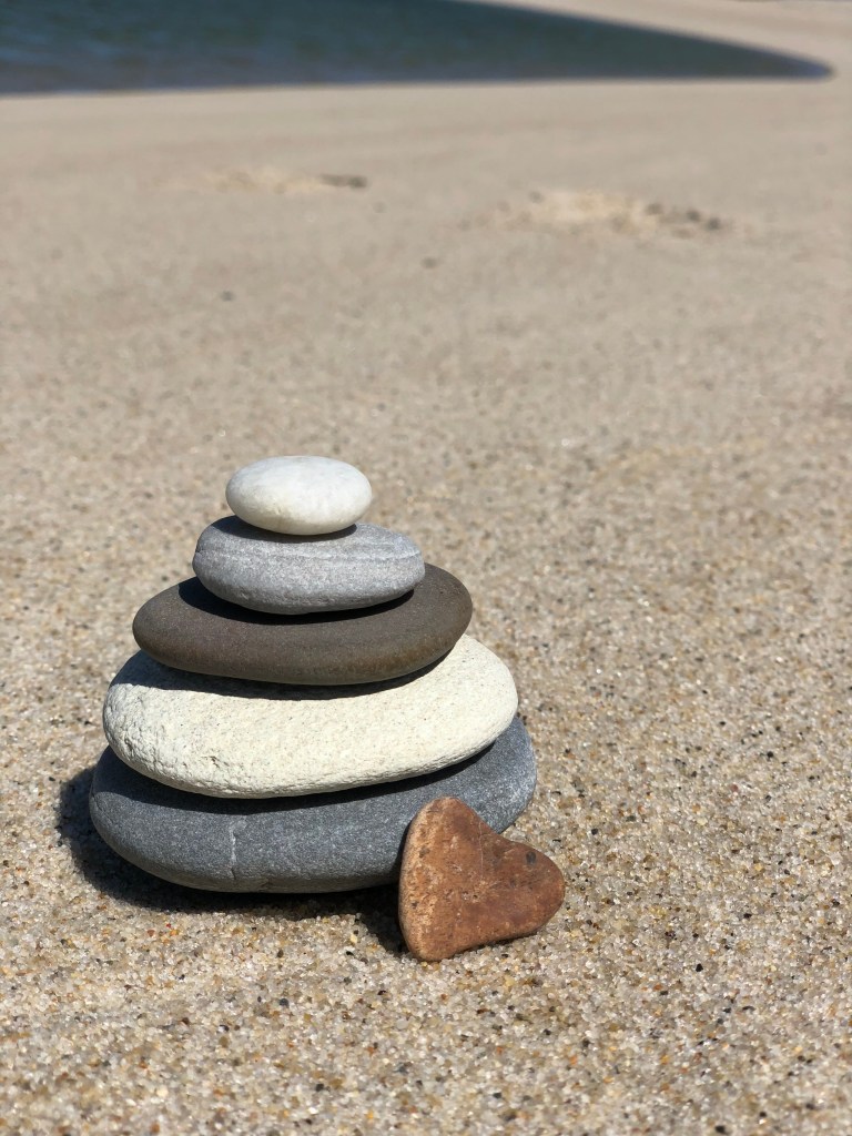 A stack of smooth stones arranged in a pyramid shape on sandy beach, accompanied by a heart-shaped stone.