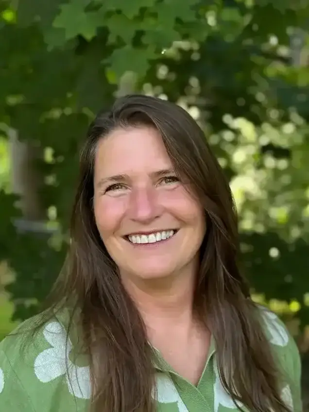 Smiling woman with long brown hair wearing a green floral shirt, standing outdoors in front of greenery.