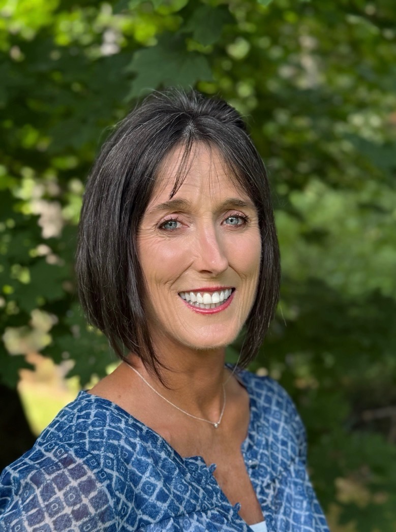 Smiling woman with shoulder-length dark hair, wearing a blue patterned blouse, in front of a blurred green background.