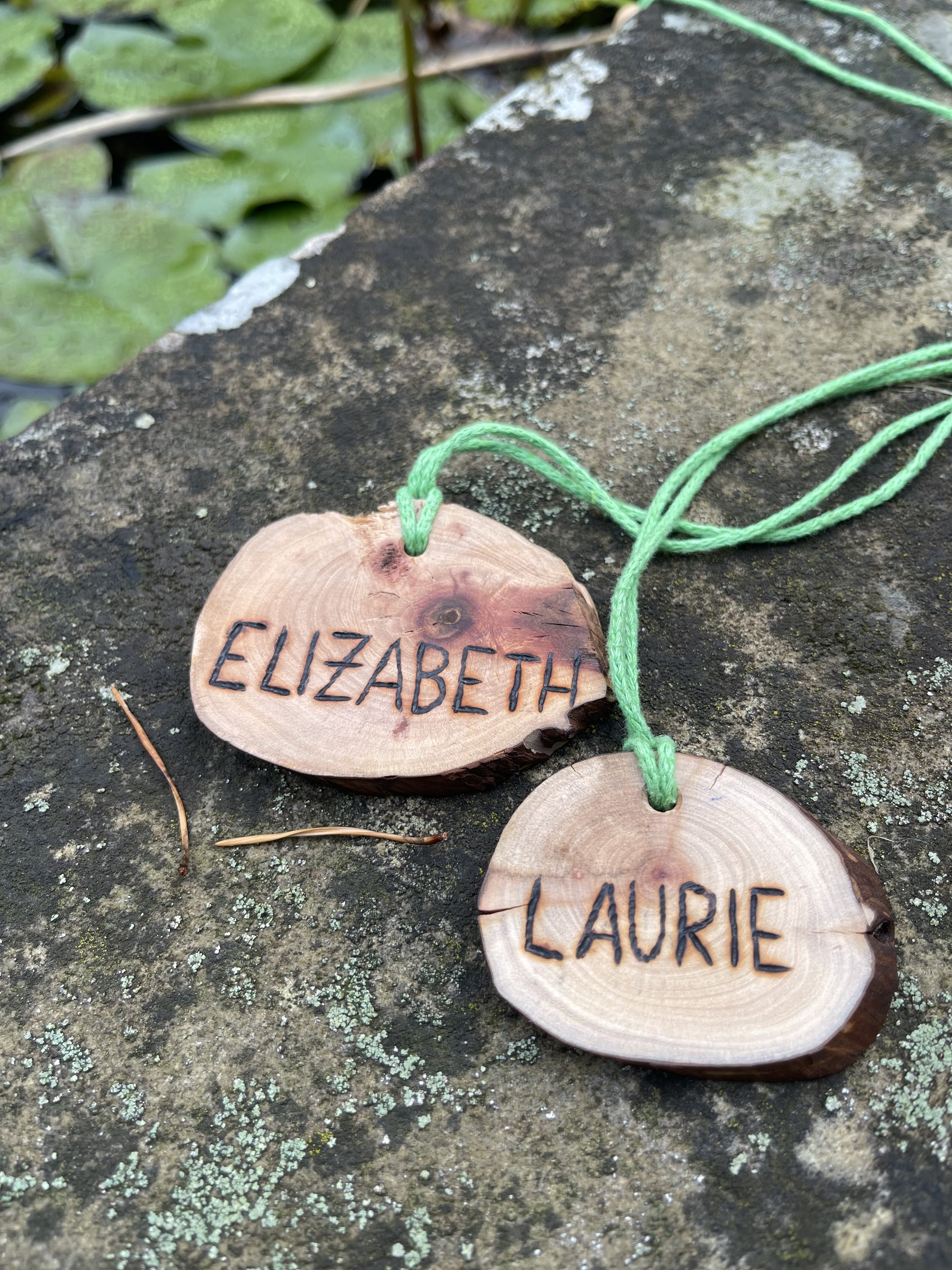 Two wooden pendants with the names 'ELIZABETH' and 'LAURIE' engraved, tied with green string, resting on a stone surface near a pond with lily pads.